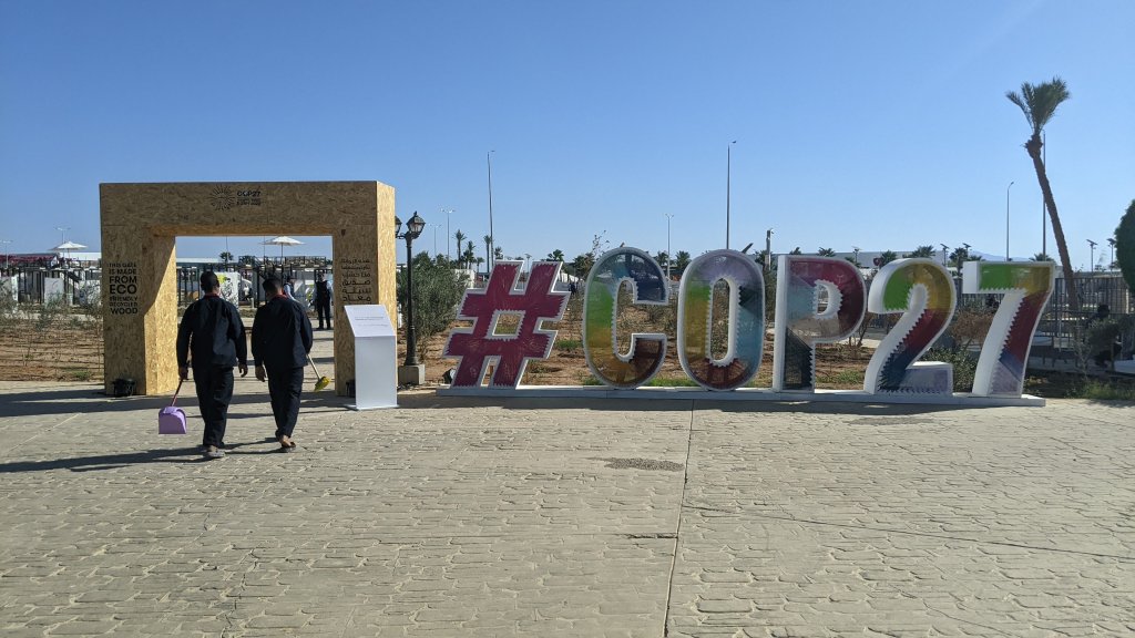 two men in dark blue uniforms holding brooms walk away from the camera next to a large multicolored sign that reads #COP27
